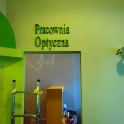 Ściana pomalowana na jasnozielono z napisem 'Pracownia Optyczna' nad wejściem. Widoczna drabina i narzędzia malarskie, sugerujące świeżo zakończone prace.