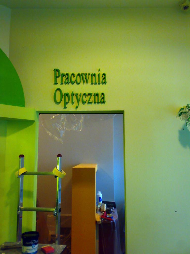 Ściana pomalowana na jasnozielono z napisem 'Pracownia Optyczna' nad wejściem. Widoczna drabina i narzędzia malarskie, sugerujące świeżo zakończone prace.