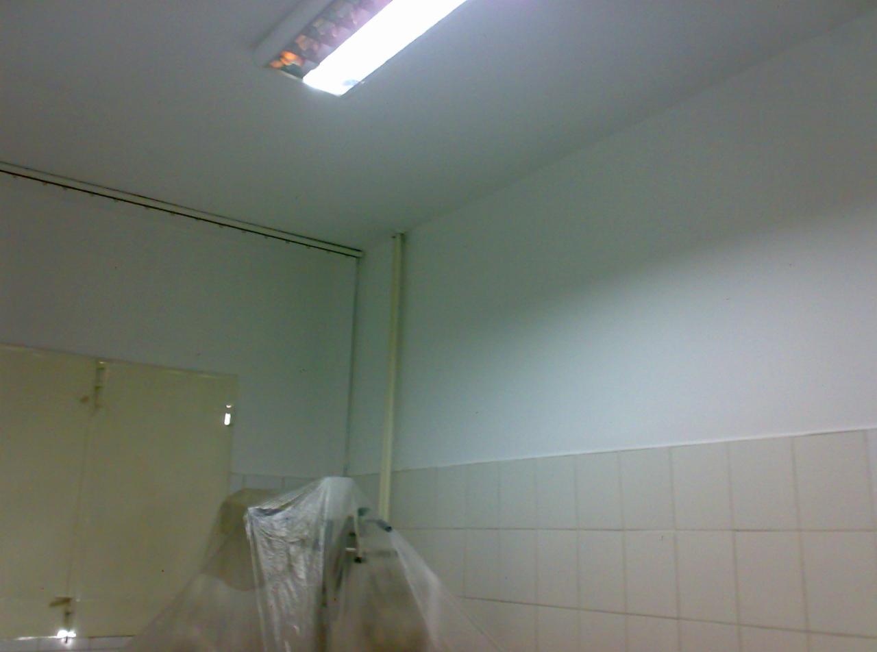 Białe ściany po malowaniu w pomieszczeniu z płytkami. Widoczna lampa sufitowa i zabezpieczone folią sprzęty. Świeżo odnowione wnętrze.