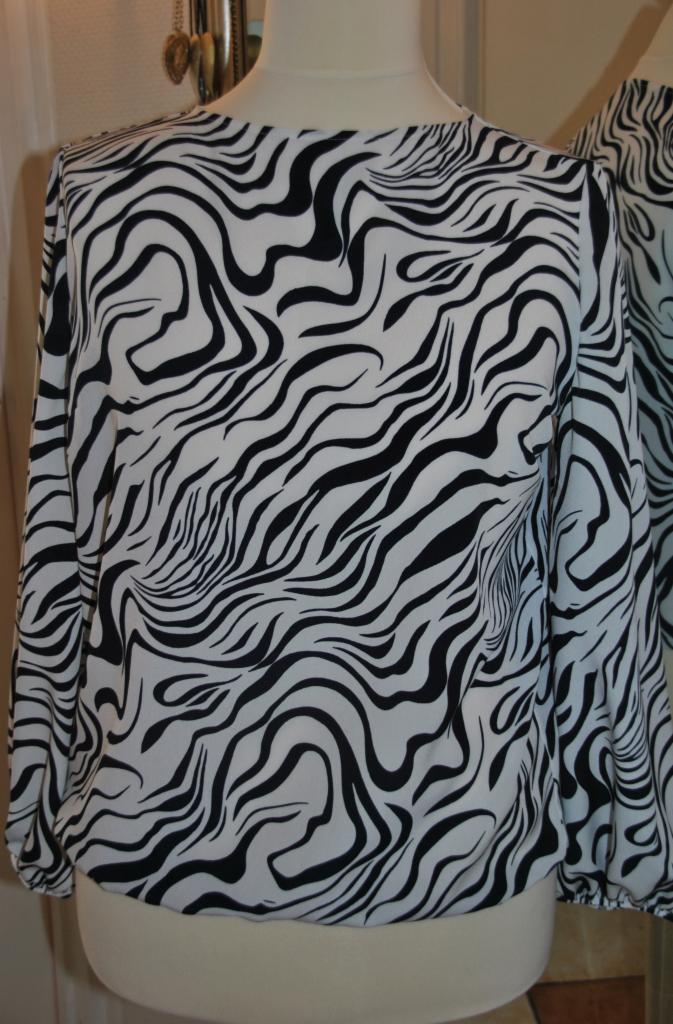 Bluzka damska z długim rękawem w biało-czarny wzór zebry, prezentowana na manekinie. Widoczne detale marszczeń na dole i przy mankietach, odbicie w lustrze w tle.