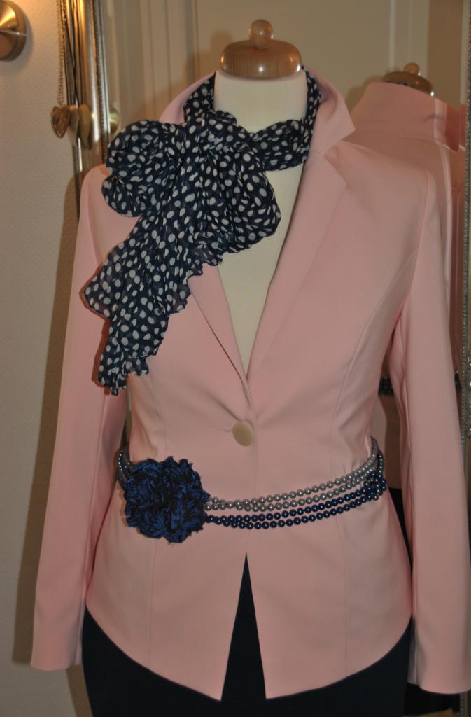 Różowy, dopasowany żakiet damski na manekinie, zdobiony wzorzystą apaszką, kwiatem i perłowym paskiem, odbijający się w lustrze w tle. Elegancka stylizacja.