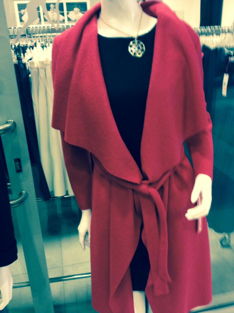 Czerwony płaszcz damski z wiązaniem w talii na manekinie, z czarną sukienką i złotym naszyjnikiem. W tle wieszaki z ubraniami w sklepie odzieżowym.