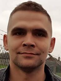 Portret mężczyzny o krótkich włosach w czarnej kurtce, na rozmytym tle z elementami zabudowy. Ujęcie od przodu, skupione na twarzy.