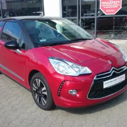 Czerwony Citroen DS3 z czarnym dachem, zaparkowany przed salonem samochodowym z widocznymi odbiciami w szybie. Dodatkowo widoczny biały samochód w tle.