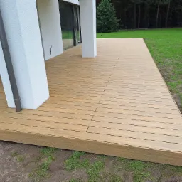 Nowoczesny taras z desek kompozytowych w kolorze jasnego drewna, przylegający do białego budynku z dużymi oknami, widok na trawnik i drzewa w tle.