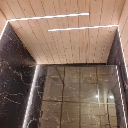 Nowoczesna łazienka z kabiną prysznicową: ściany wyłożone czarnym marmurem z białymi żyłami, sufit z jasnego drewna z wpuszczonymi liniowymi lampami LED, szklana kabina prysznicowa odbijająca wnętrze.