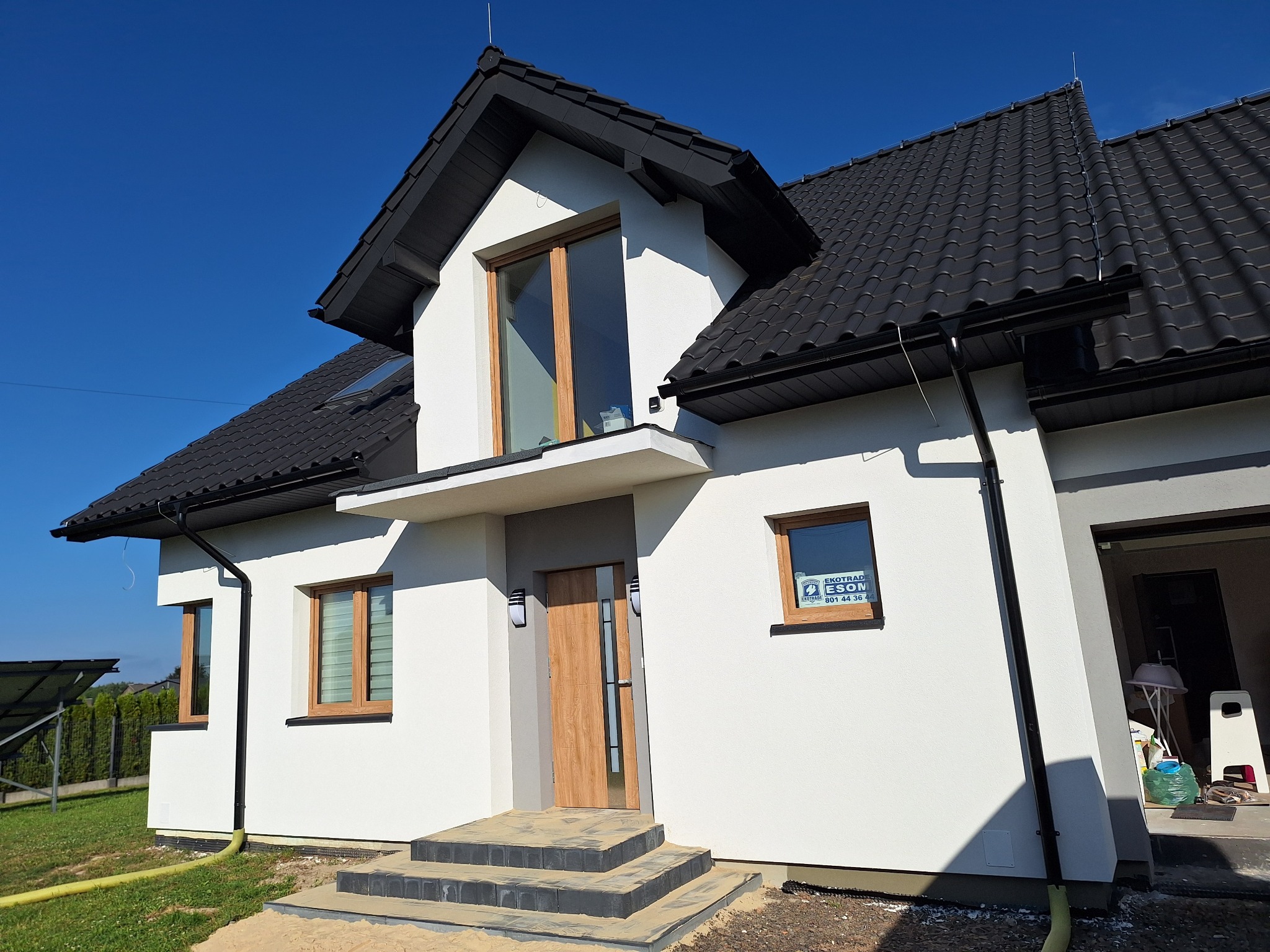 Nowoczesny dom jednorodzinny z białą elewacją, czarnym dachem i drewnianymi oknami w słoneczny dzień. Widoczne schody wejściowe wyłożone kostką i fragment instalacji fotowoltaicznej w tle.