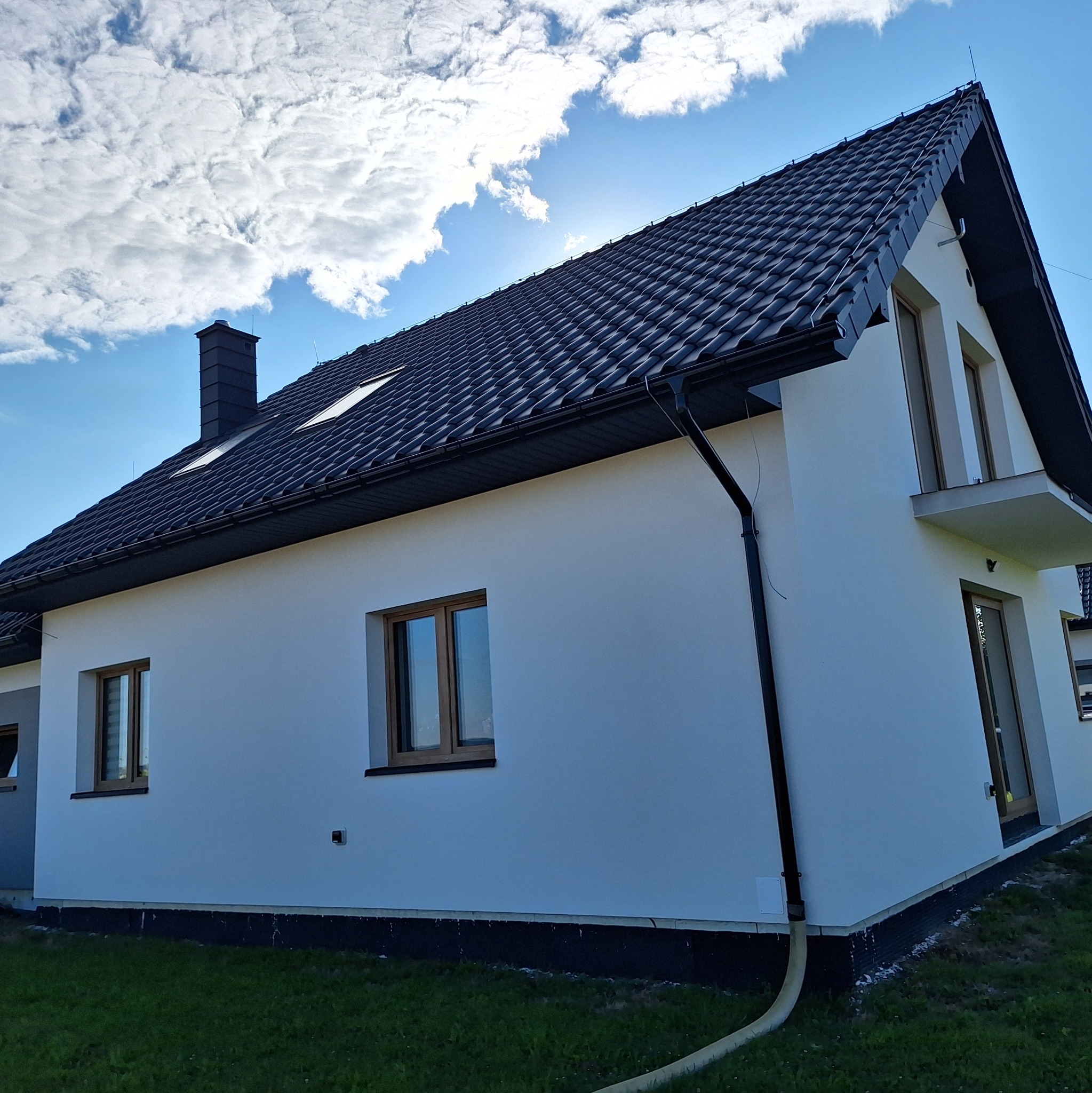 Nowoczesny dom jednorodzinny z białą elewacją, czarnym dachem z dachówki i brązowymi oknami, sfotografowany w słoneczny dzień z widocznymi chmurami na niebie.