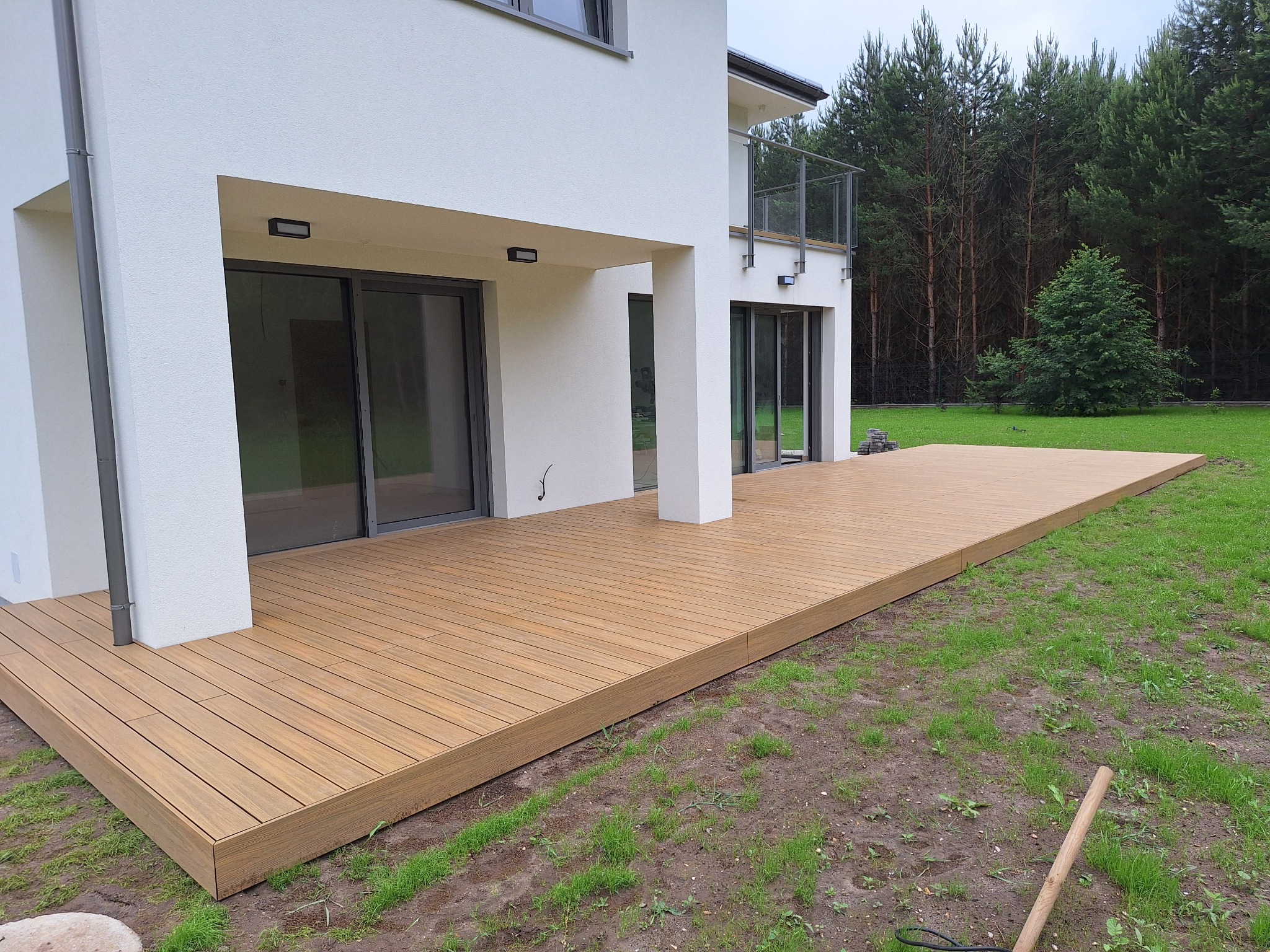 Gotowa drewniana deska tarasowa przylegająca do białego domu z dużymi przesuwnymi drzwiami, widok na trawnik i las w tle.