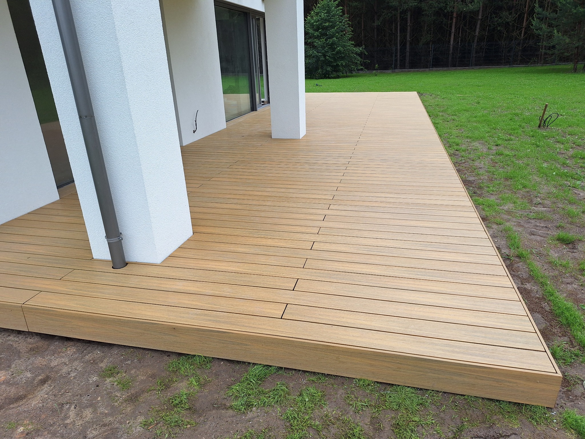 Nowoczesny taras z desek kompozytowych w kolorze jasnego drewna, przylegający do białego budynku z dużymi oknami, widok na trawnik i drzewa w tle.