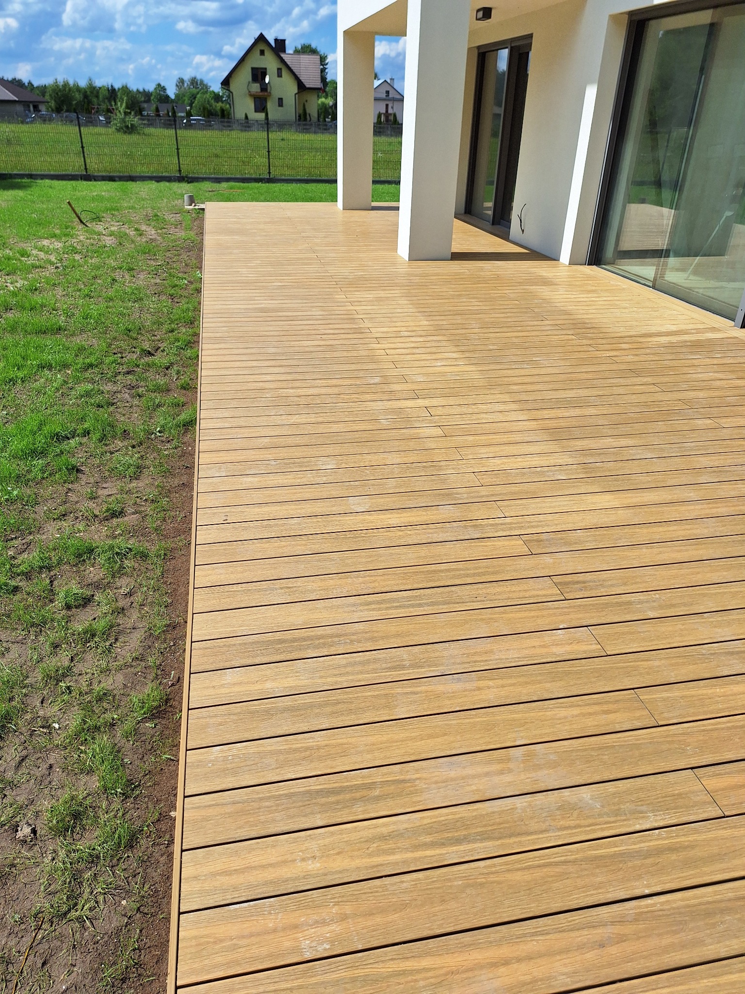 Nowoczesny taras z desek kompozytowych w Mniowie, przylegający do białego budynku z dużymi oknami, widok na trawnik i dom w tle.