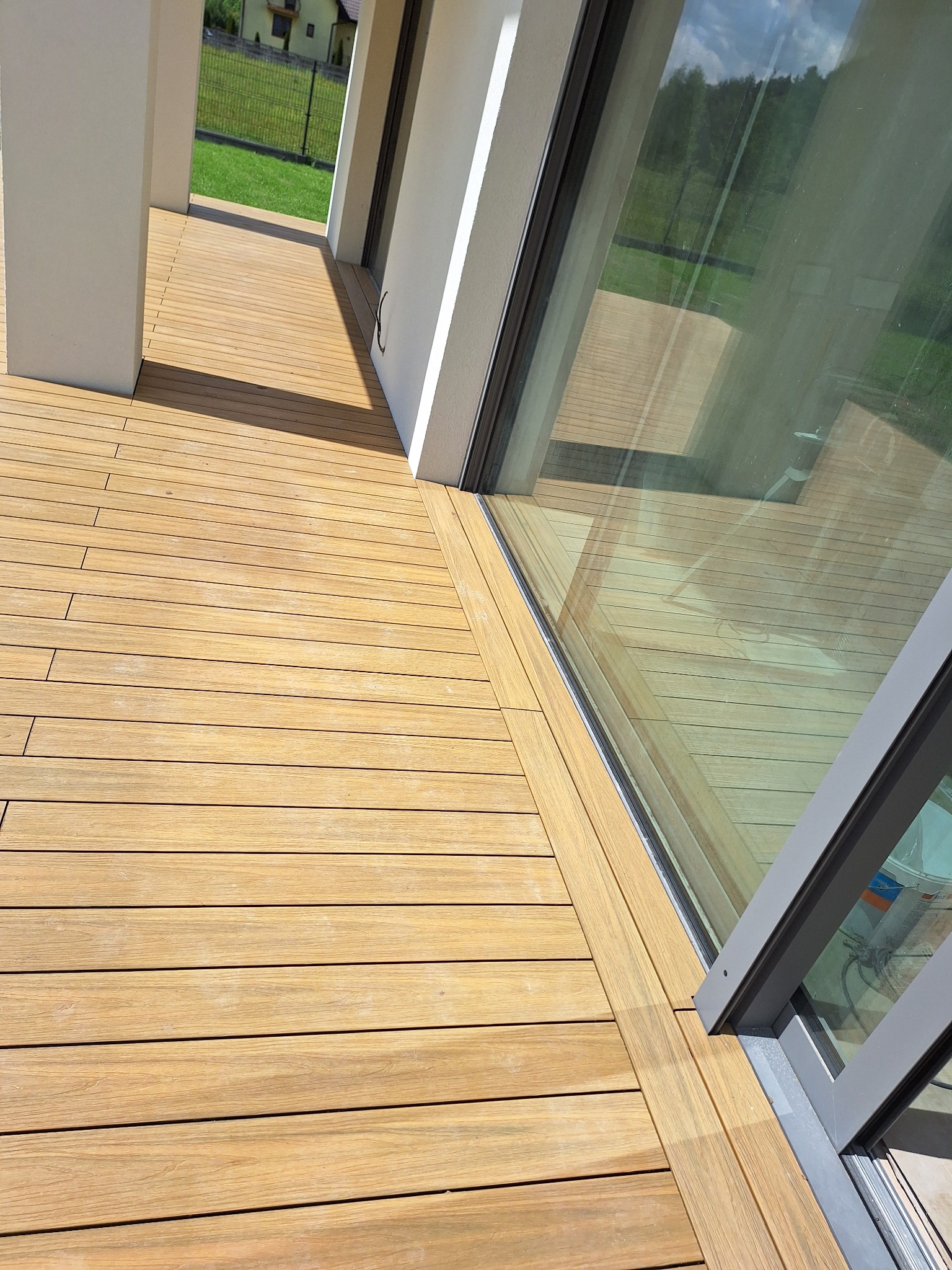 Jasna, drewniana podłoga tarasu z kompozytu ułożona wzdłuż dużego okna tarasowego z szarymi ramami, widok na trawnik i dom w tle.