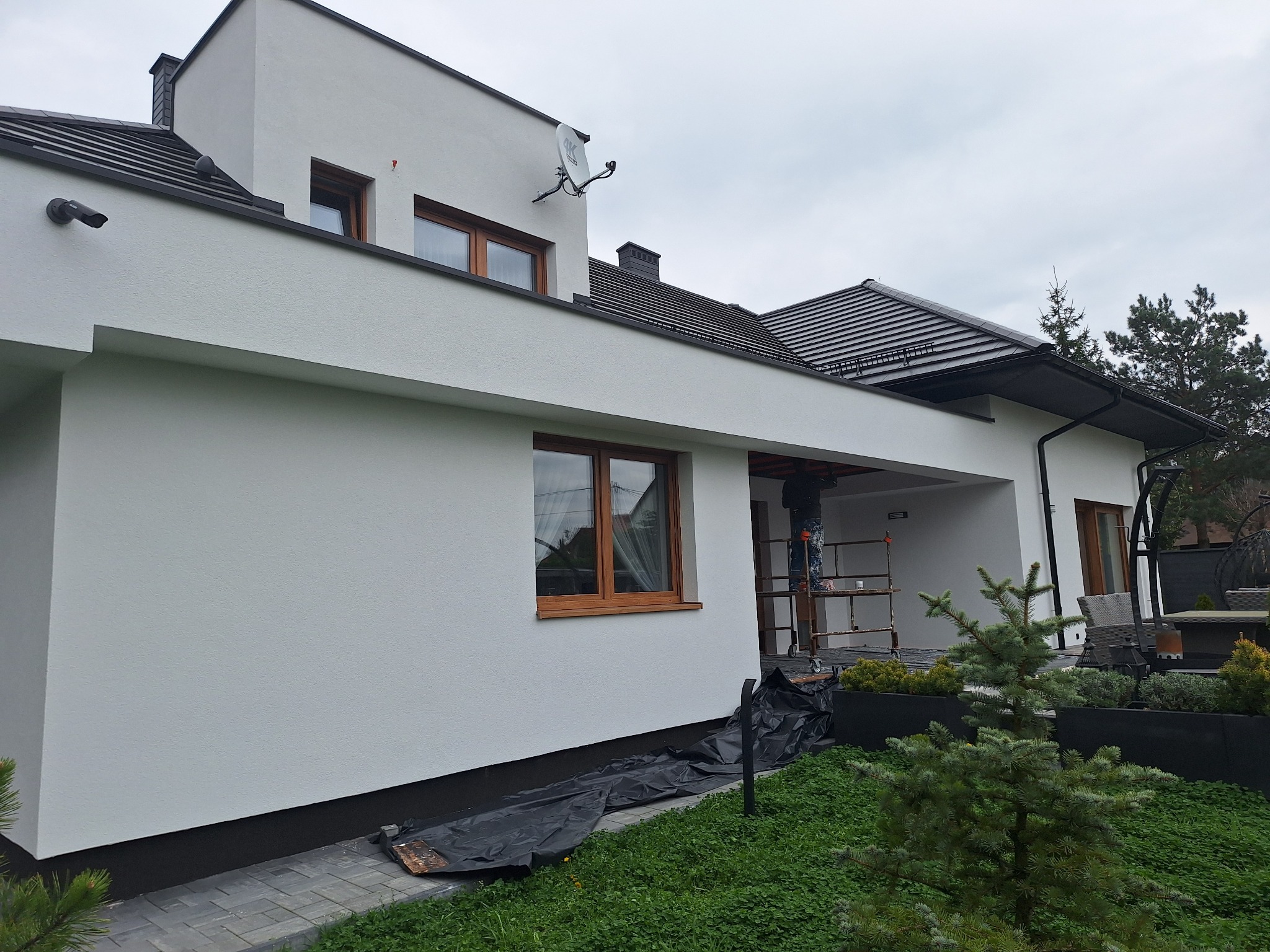 Nowoczesny dom jednorodzinny z białą elewacją, drewnianymi oknami i ciemną dachówką, w trakcie prac wykończeniowych, widoczne rusztowanie i zabezpieczenia na trawniku.