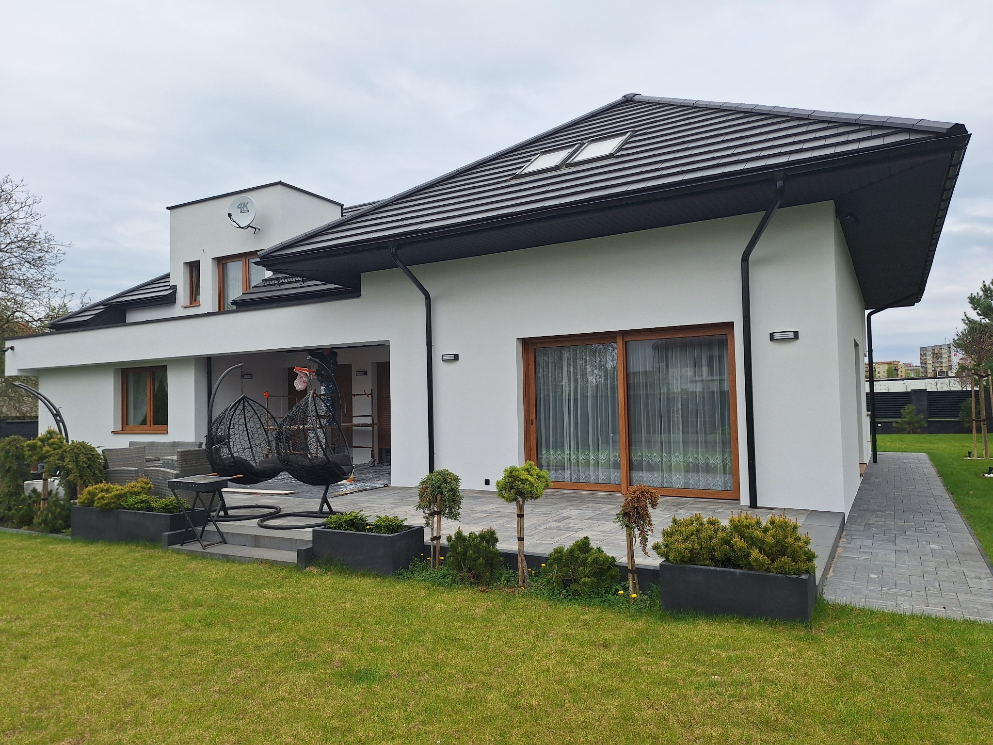 Nowoczesny dom jednorodzinny z białą elewacją, czarnym dachem z oknami dachowymi, tarasem z wiszącymi fotelami i zadbanym trawnikiem z ozdobnymi roślinami w donicach.