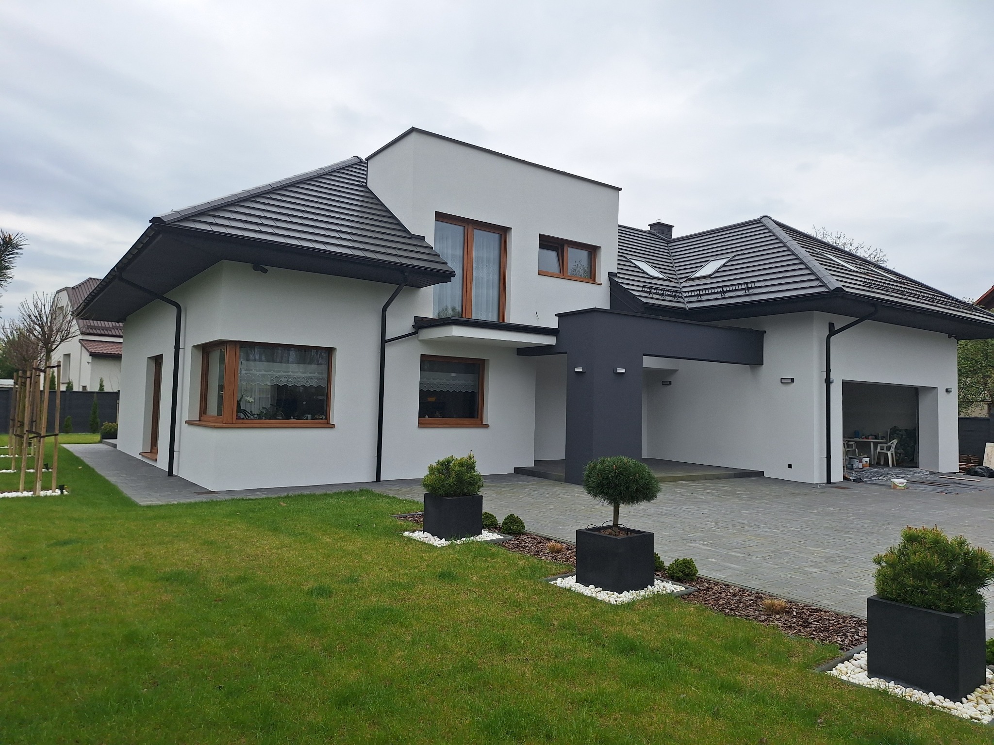 Nowoczesny dom jednorodzinny z białymi ścianami i grafitowym akcentem nad wjazdem do garażu, ciemna dachówka, zadbany trawnik z ozdobnymi krzewami w donicach.