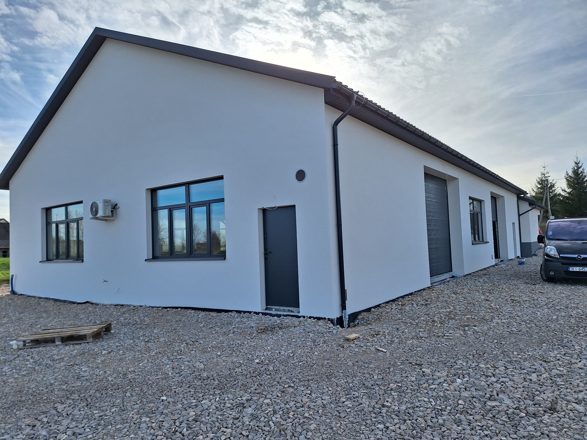 Nowoczesny budynek z jasną elewacją, grafitowymi oknami, drzwiami i elementami wykończeniowymi, widoczny klimatyzator na ścianie, teren wokół wysypany żwirem, częściowo widoczny samochód.