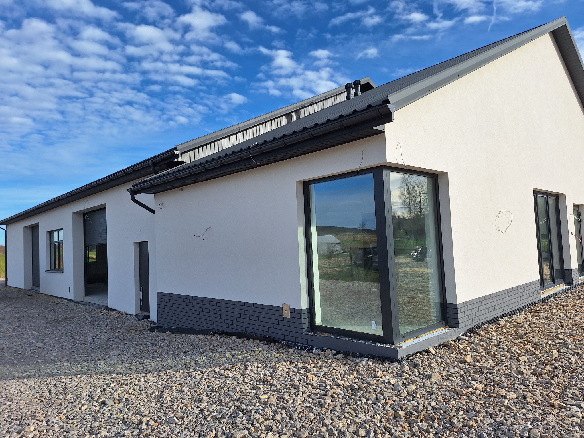Nowoczesny budynek jednokondygnacyjny z białymi ścianami i szarą podmurówką z cegły, duże okna z czarnymi ramami, szary dach, teren wokół wysypany żwirem, błękitne niebo z chmurami w tle.
