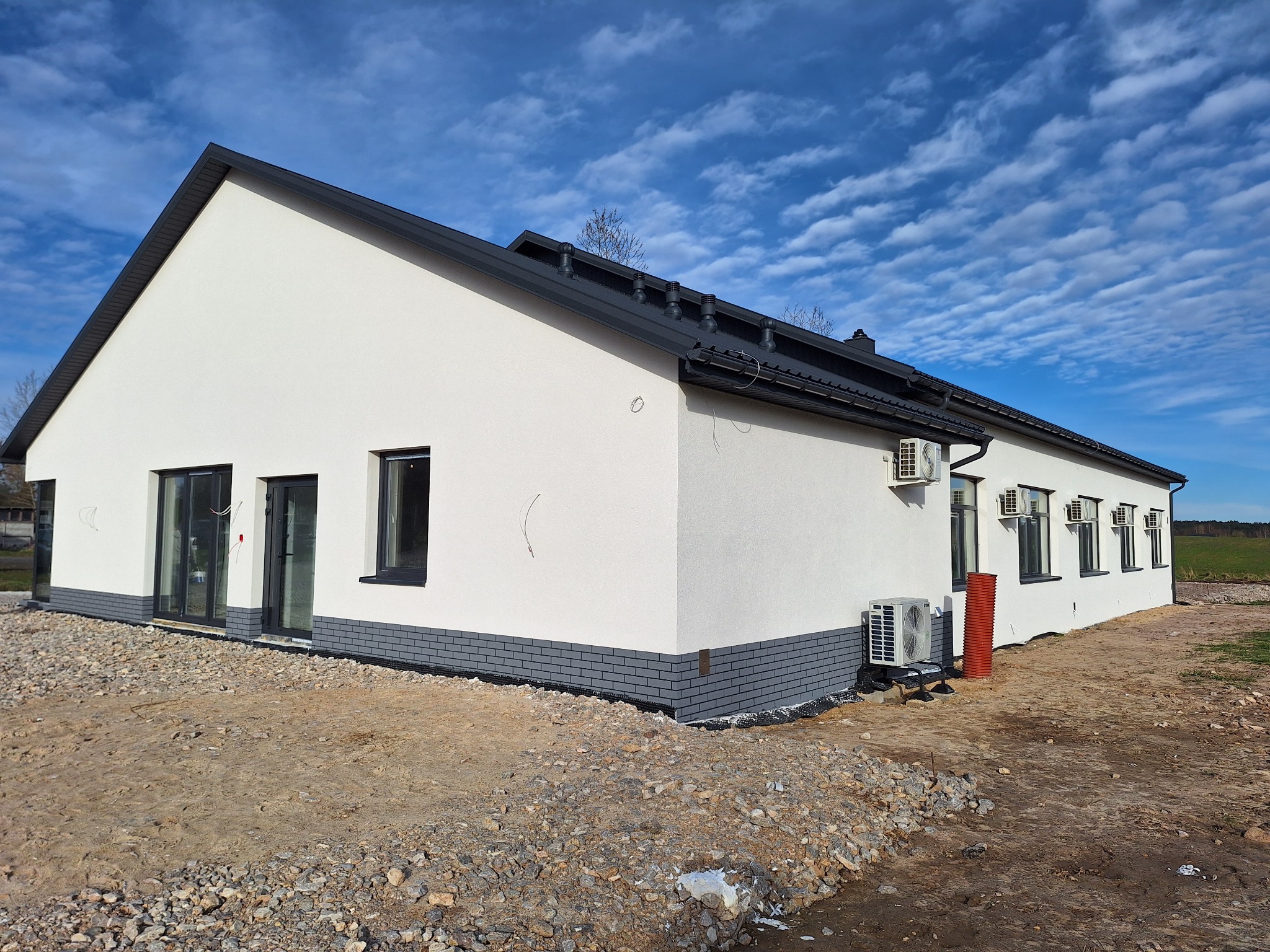 Nowoczesny, parterowy dom z białymi ścianami i ciemną stolarką okienną, wykończony szarą cegłą klinkierową u podstawy, widoczne jednostki klimatyzacyjne na elewacji bocznej, teren wokół domu...