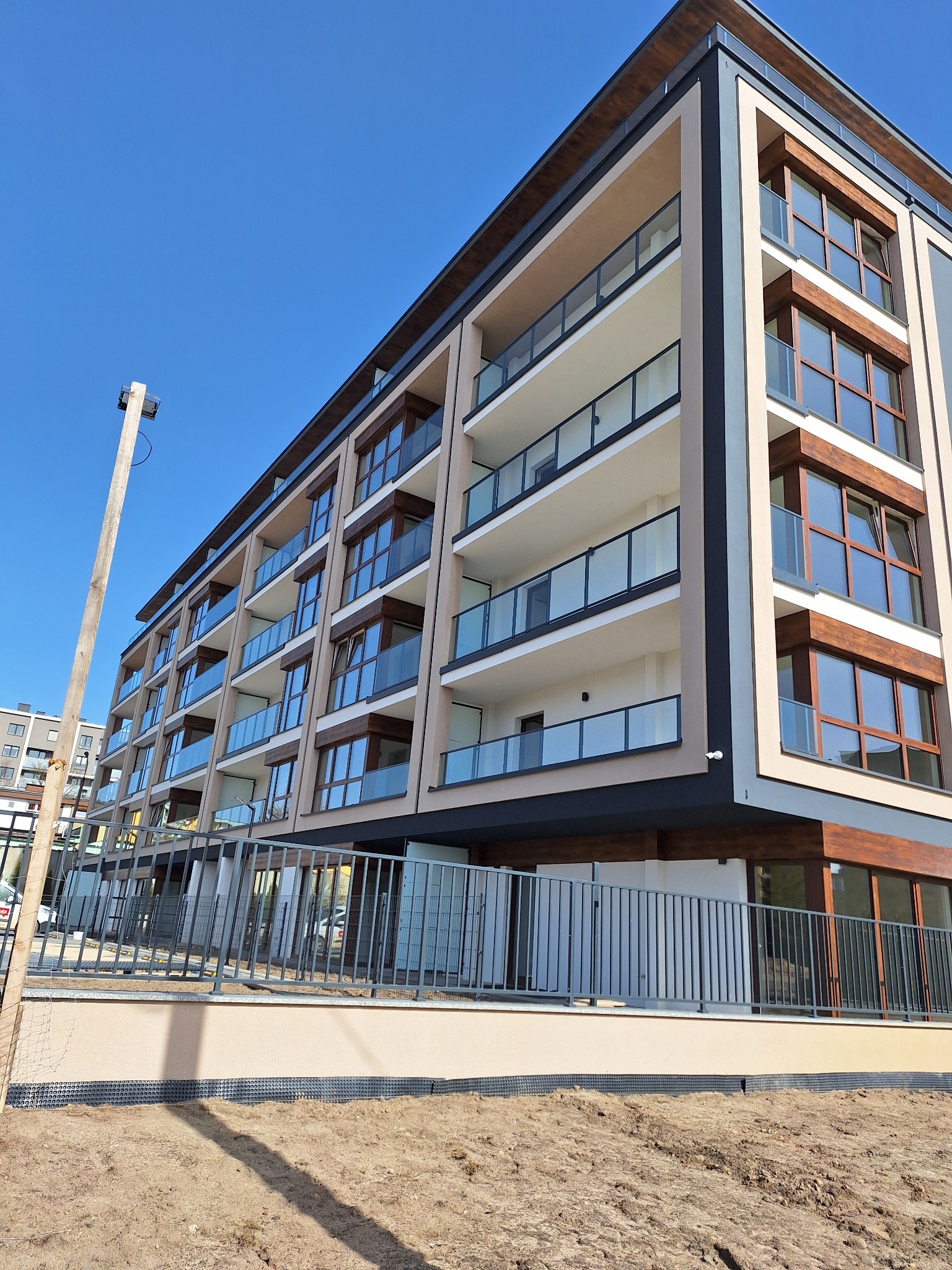 Nowoczesny budynek wielorodzinny z balkonami o szklanych balustradach, wykończony w beżach i brązach, widziany pod kątem od dołu w słoneczny dzień. Teren wokół budynku przygotowany...