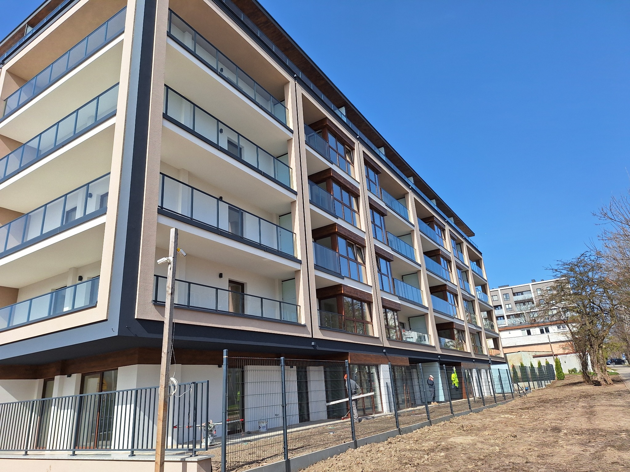 Nowoczesny budynek wielorodzinny z balkonami o szklanych balustradach, świeżo po budowie, widoczny z poziomu gruntu, z fragmentem ogrodzenia i przygotowanym terenem.