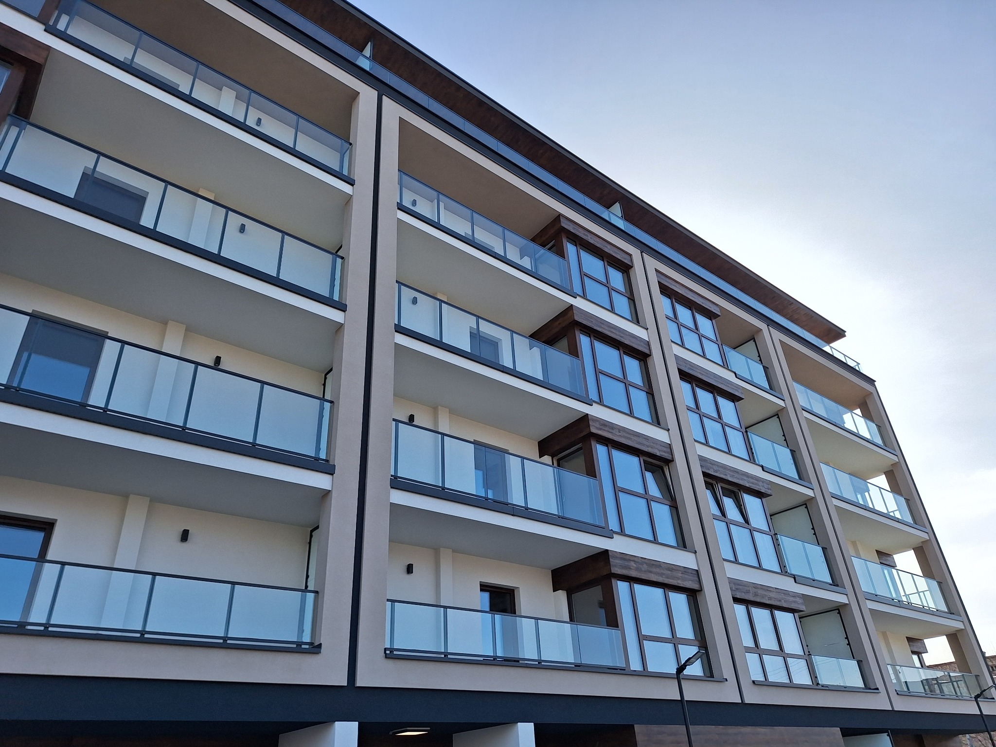 Nowoczesny budynek wielorodzinny z beżową elewacją, szklanymi balkonami i drewnianymi ramami okiennymi sfotografowany pod kątem od dołu.