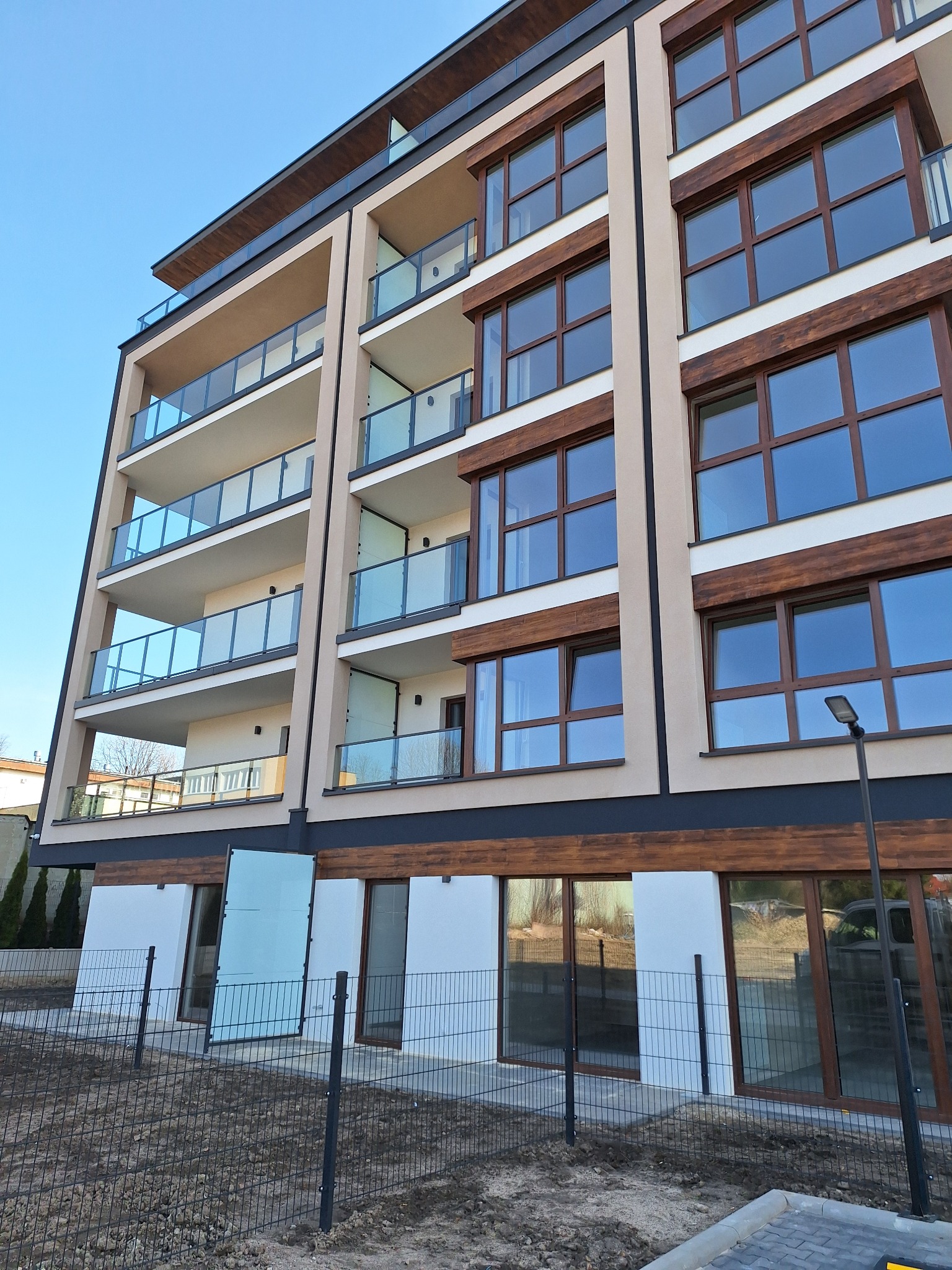 Nowoczesny budynek wielorodzinny z beżową elewacją, drewnopodobnymi elementami wykończeniowymi okien i balkonami z przezroczystymi balustradami, ogrodzony metalową siatką, w słoneczny dzień.