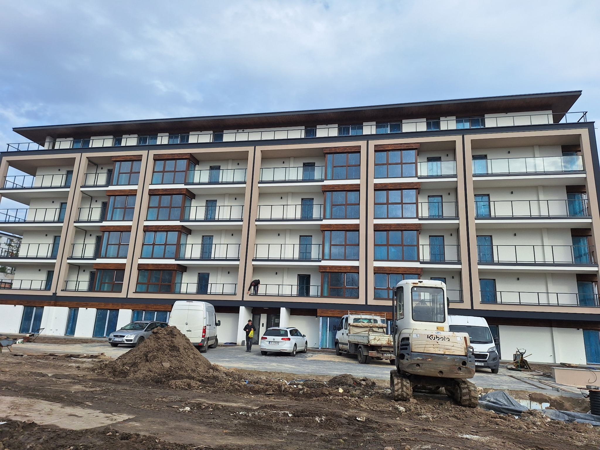 Wielopiętrowy budynek z balkonami w trakcie wykończenia elewacji, z widocznym sprzętem budowlanym i materiałami na placu budowy.