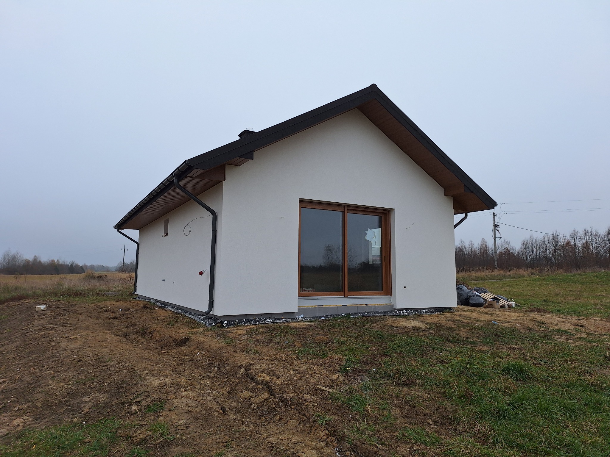 Nowoczesny, parterowy dom z białymi ścianami i brązowymi ramami okiennymi, z czarną dachówką i rynnami, na działce w trakcie zagospodarowywania, widok z zewnątrz w pochmurny dzień.