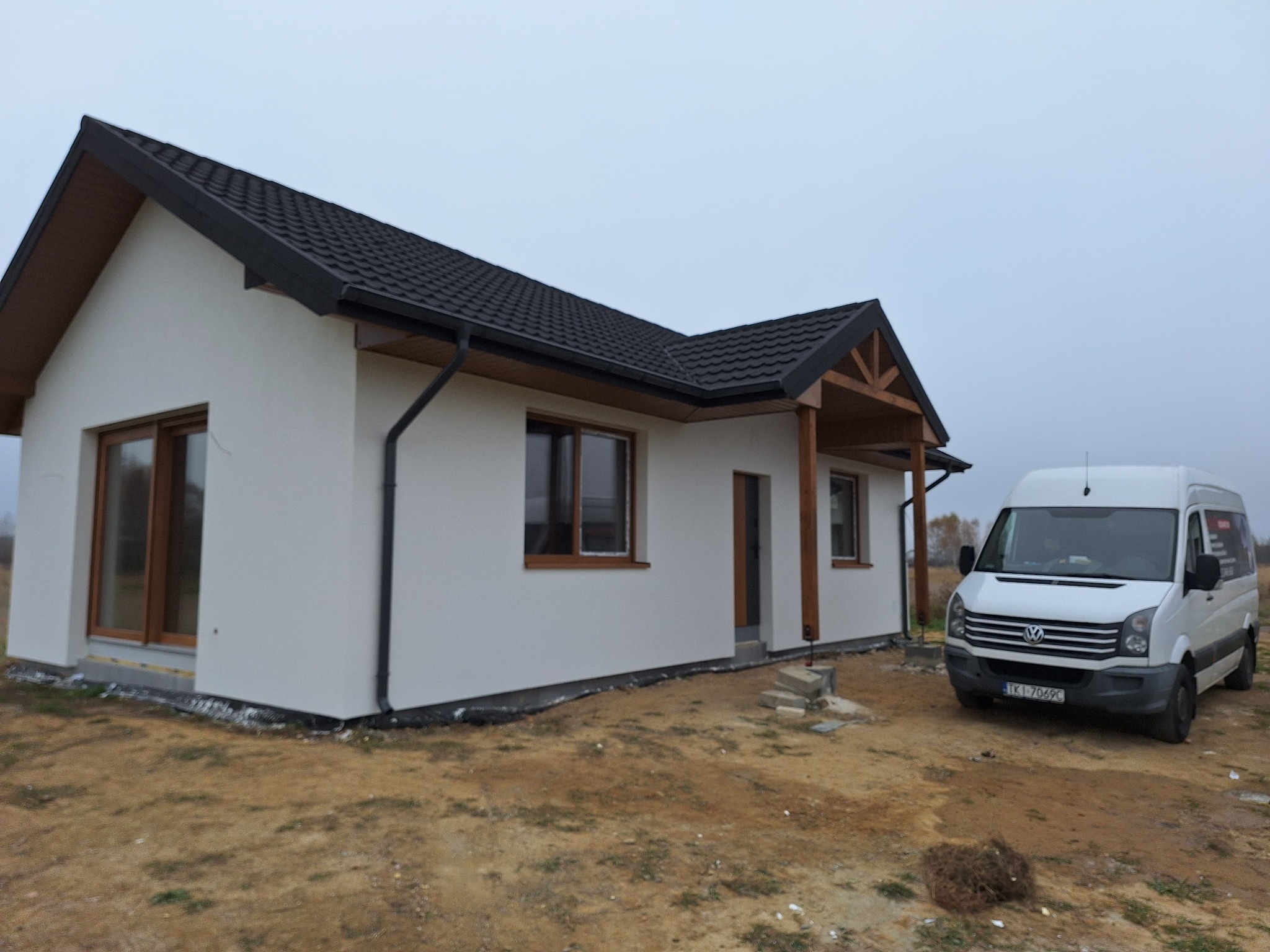 Dom jednorodzinny z białą elewacją i ciemnym dachem, widoczny z boku, z zaparkowanym białym vanem. Teren wokół domu częściowo przygotowany, z widocznymi bloczkami fundamentowymi.