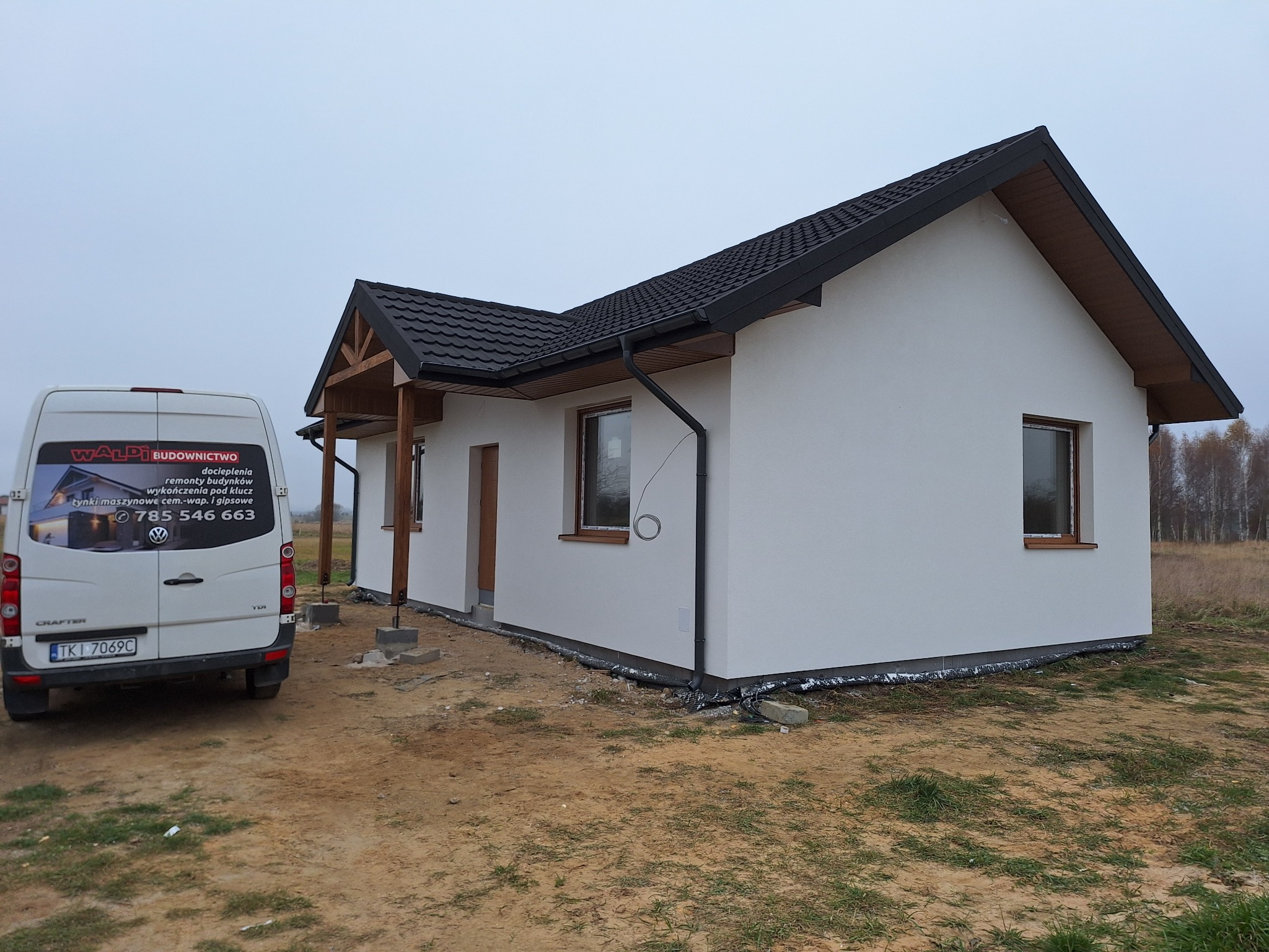 Nowo wybudowany, otynkowany dom jednorodzinny z czarnym dachem i drewnianymi elementami wykończeniowymi, widziany z boku, z zaparkowanym białym vanem z reklamą firmy budowlanej w tle.