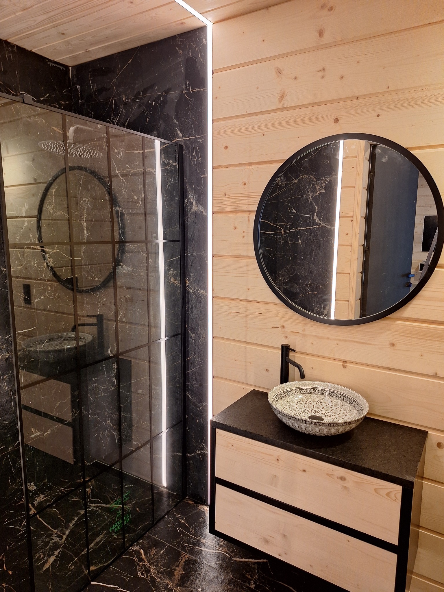Nowoczesna łazienka z kabiną prysznicową wykończoną czarnym marmurem, drewnianymi panelami ściennymi, okrągłym lustrem i ozdobną umywalką nablatową.