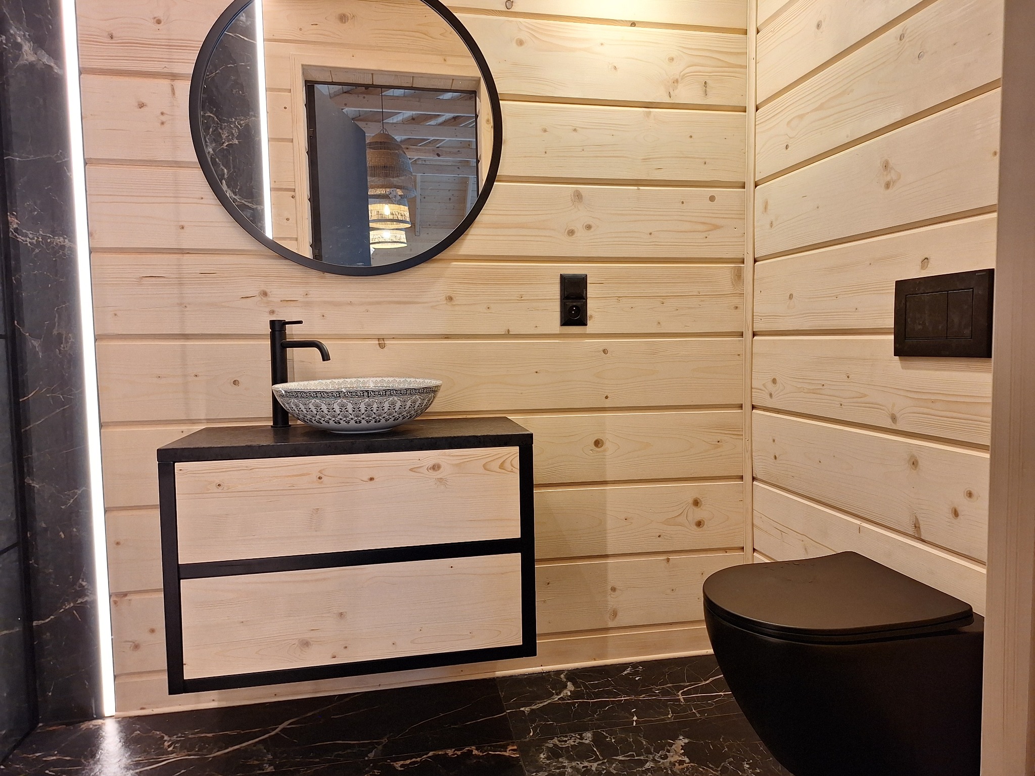 Nowoczesna łazienka w Mniowie z drewnianą boazerią, czarną armaturą i marmurową podłogą. Okrągłe lustro z podświetleniem LED odbija designerską lampę. Uwagę zwraca umywalka w stylu boho.