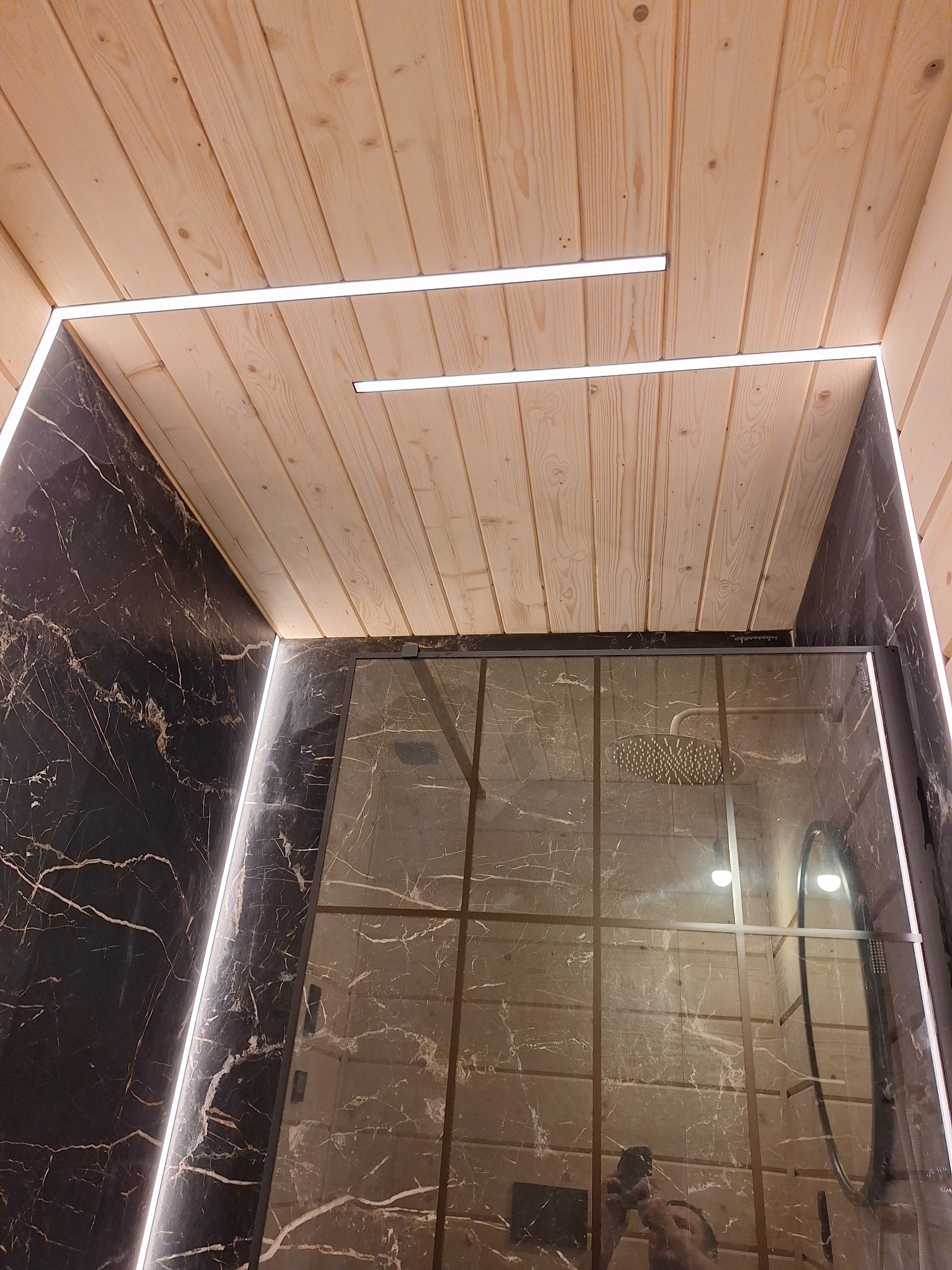 Nowoczesna łazienka z kabiną prysznicową: ściany wyłożone czarnym marmurem z białymi żyłami, sufit z jasnego drewna z wpuszczonymi liniowymi lampami LED, szklana kabina prysznicowa odbijająca wnętrze.