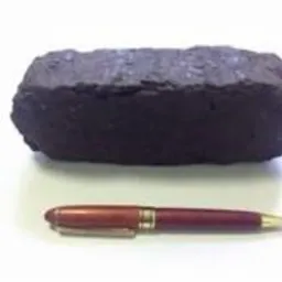 Bryła ciemnego brykietu węglowego obok długopisu na białym tle, ukazująca jego wielkość i strukturę. Widoczne nierówności i pęknięcia na powierzchni brykietu.