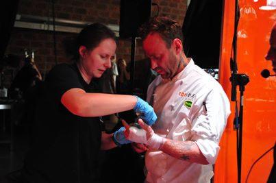 Instruktor z Ostrowic demonstruje bandażowanie ręki na manekinie podczas kursu pierwszej pomocy. Uczestnik w stroju kucharza, zbliżenie na dłonie.