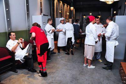 Szkolenie z pierwszej pomocy dla kucharzy: instruktor w czerwonym kombinezonie demonstruje techniki na siedzącym kucharzu. Grupa obserwuje w tle.