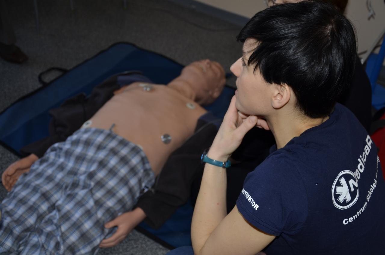 Instruktorka z ręką na brodzie obserwuje manekina podczas kursu pierwszej pomocy. Widoczny napis 'Instruktor' na koszulce i logo centrum szkoleniowego.