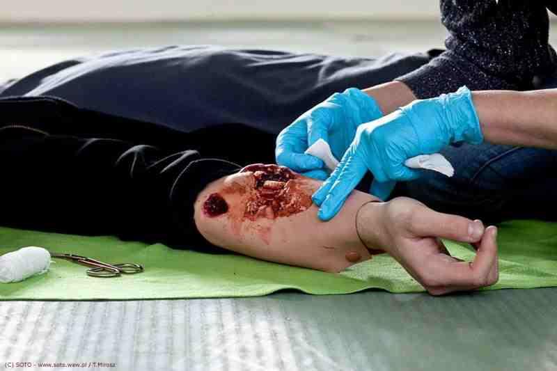 Opatrywanie rany na ramieniu manekina podczas kursu pierwszej pomocy. Widoczne ręce w niebieskich rękawiczkach i narzędzia medyczne.