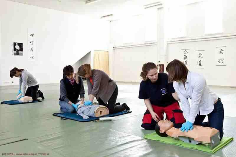 Szkolenie z pierwszej pomocy: grupa osób ćwiczy resuscytację na manekinach w jasnej sali z napisami w języku orientalnym na ścianie.