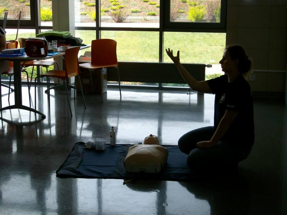 Instruktorka na kolanach demonstruje resuscytację na manekinie podczas kursu pierwszej pomocy. Widoczne okno i wyposażenie sali.