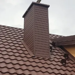 Brązowy dach z blachodachówki z obudowanym kominem z blachy trapezowej, zabezpieczenie przeciwśniegowe na połaci dachu.