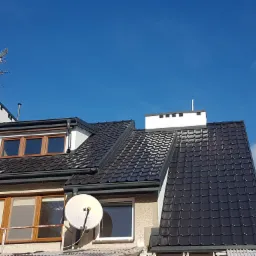 Widok z dołu na czarny dach z dachówki ceramicznej, białe kominy, okna z brązowymi ramami, antenę telewizyjną i satelitarną na tle błękitnego nieba z lekkimi chmurami.