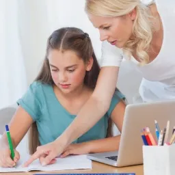 Dziewczynka pisze w zeszycie pod okiem nauczycielki, laptop i przybory na biurku, jasne wnętrze.