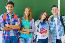 Grupa uśmiechniętych nastolatków z plecakami i książkami, stojąca na tle zielonej tablicy szkolnej.