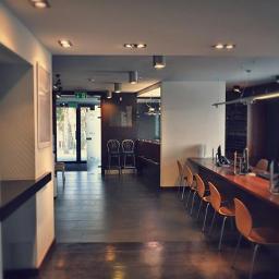 CCTEL Sp. z o.o. Sp. k. - Nowoczesne wnętrze biurowe z długim drewnianym blatem, pomarańczowymi krzesłami, komputerami i widokiem na przeszklone drzwi wejściowe z zielonym znakiem ewakuacyjnym.