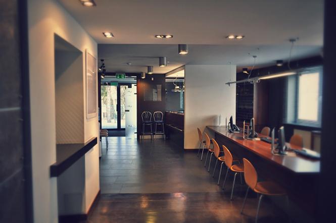 Nowoczesne wnętrze biurowe z długim drewnianym blatem, pomarańczowymi krzesłami, komputerami i widokiem na przeszklone drzwi wejściowe z zielonym znakiem ewakuacyjnym.