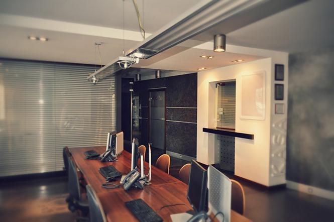 Nowoczesne biuro z długim, drewnianym stołem konferencyjnym, wyposażonym w komputery i telefony stacjonarne, oświetlone listwą LED pod sufitem oraz z żaluzjami w oknach.