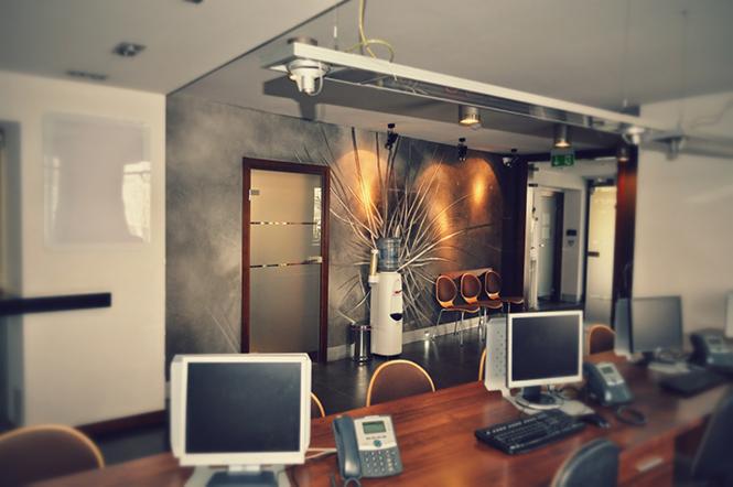 Nowoczesne biuro z długim drewnianym stołem, komputerami, telefonami, designerską ścianą z motywem roślinnym, dystrybutorem wody i trzema krzesłami w tle.