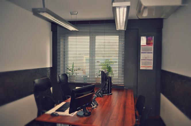 Biuro z dwoma stanowiskami komputerowymi, drewnianym biurkiem, żaluzjami w oknach i kalendarzem na ścianie.