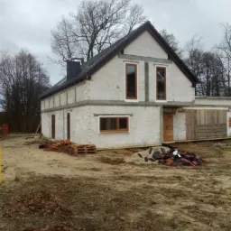 Surowy stan domu z białych bloczków, widoczne okna z brązowymi ramami, czarny komin i sterta dachówek na ziemi przed budynkiem.